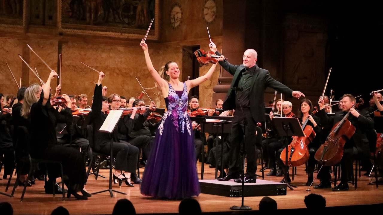 Violinist, conductor, and multiple string players in an orchestra holding batons aloft at the end of a performance.