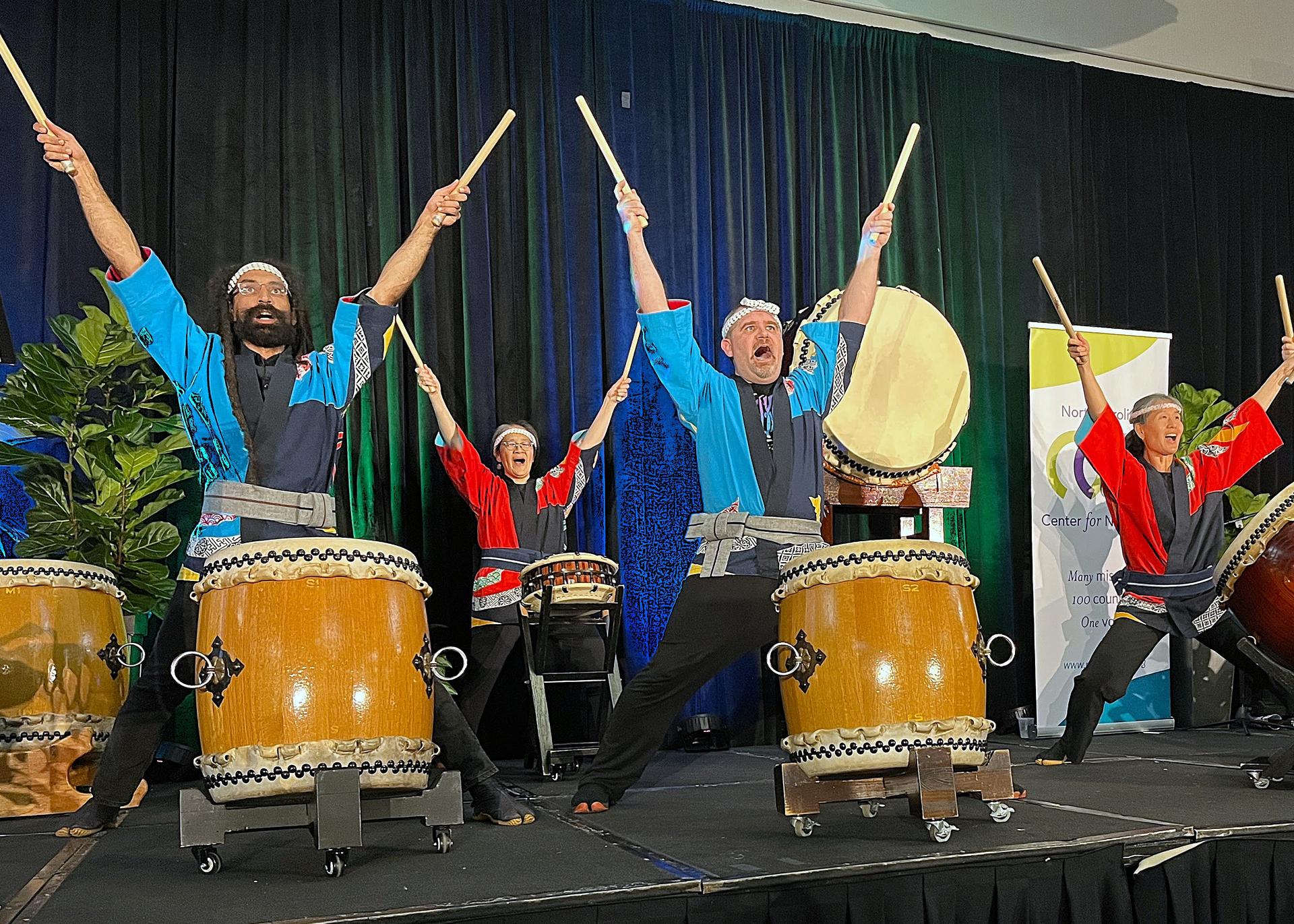 Several Triangle Taiko drummers stand behind their large drums with drumsticks raised above their heads in a performance of traditional Japanese drumming.