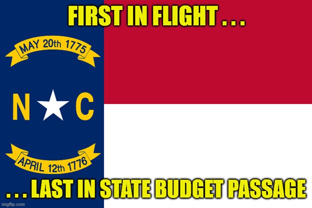 State flag of North Carolina with meme text: First in flight...last in state budget passage.